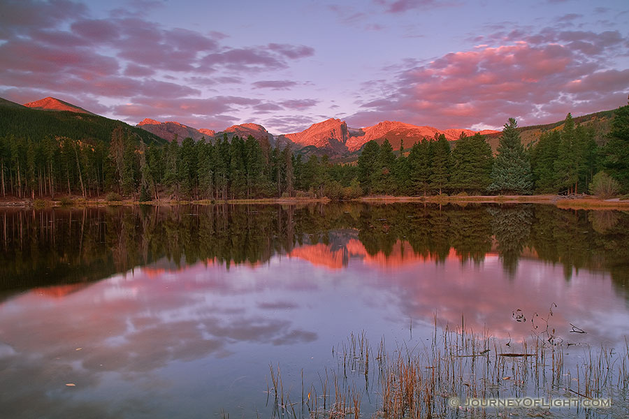 During a cool October sunrise, bright with alpenglow, the peaks of the continental divide, Powell Peak, Taylor Peak, Otis Peak, Hallett Peak, and Flattop Mountain are reflected in the calm waters of Sprague lake while underlit clouds hover overhead. - Rocky Mountain NP Photography