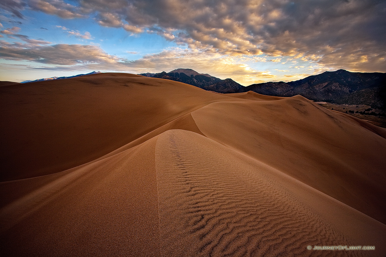 On a cool, autumn morning, silence reigned across the scenic Great Sand Dunes National Park, the quiet only occasionally broken by the sound of a breeze flowing through the dunes.  On top of one of the larger dunes, I captured the patterns of the sand as the dunes drift into one another, creating a path to the distant Sangre de Cristo Mountains.   Beyond the far peaks, the rising sun illuminates the cloud bank with a yellowish-orange tint. - Great Sand Dunes NP Picture
