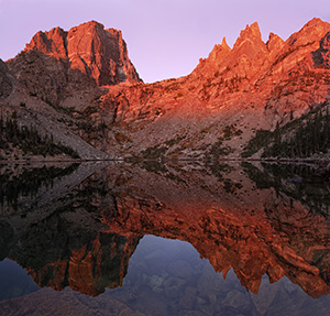 Unusually calm, Emerald Lake reflects a perfect and symmetrical image of the grand Hallet Peak and Flattop Mountain illuminated with alpenglow.  The only sounds on this quiet morning are the flow of a waterfall across the lake and the chirping of a nearby Steller's Jay. - Colorado Photograph