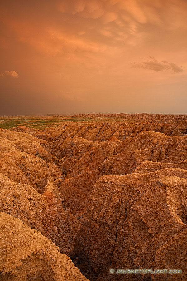 Orange hues from the last light of the setting sun illuminate storm clouds as they pass over Badlands National Park in South Dakota, dark clouds contrast with the desolate landscape. - South Dakota Photography
