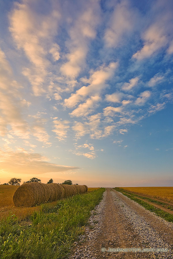 Hay bales line a country road that goes into the sunset while clouds float above. - Nebraska Photography