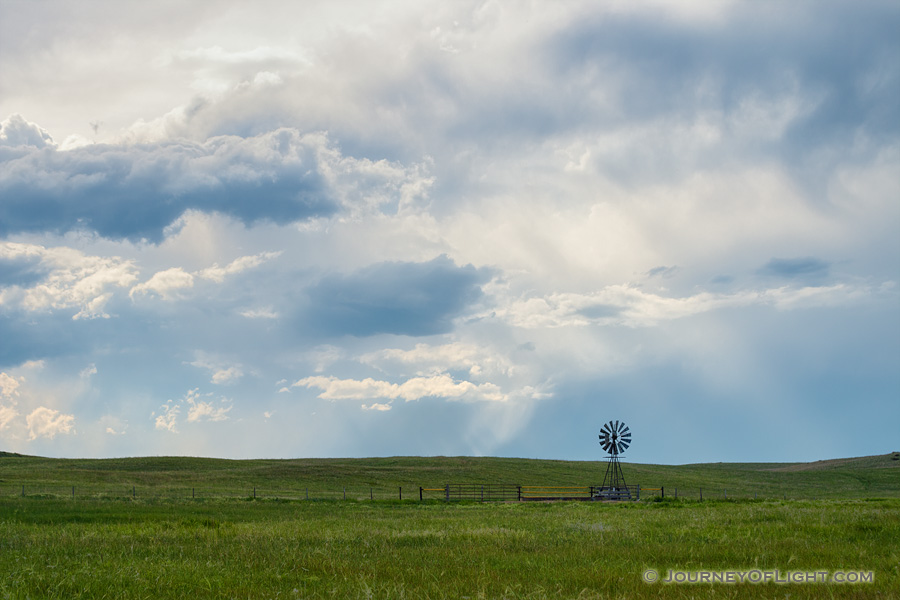 The sun peeks through a group of storm clouds illuminating a windmill deep within the Sandhills of Nebraska as storm clouds roll in the distance. - Nebraska Photography