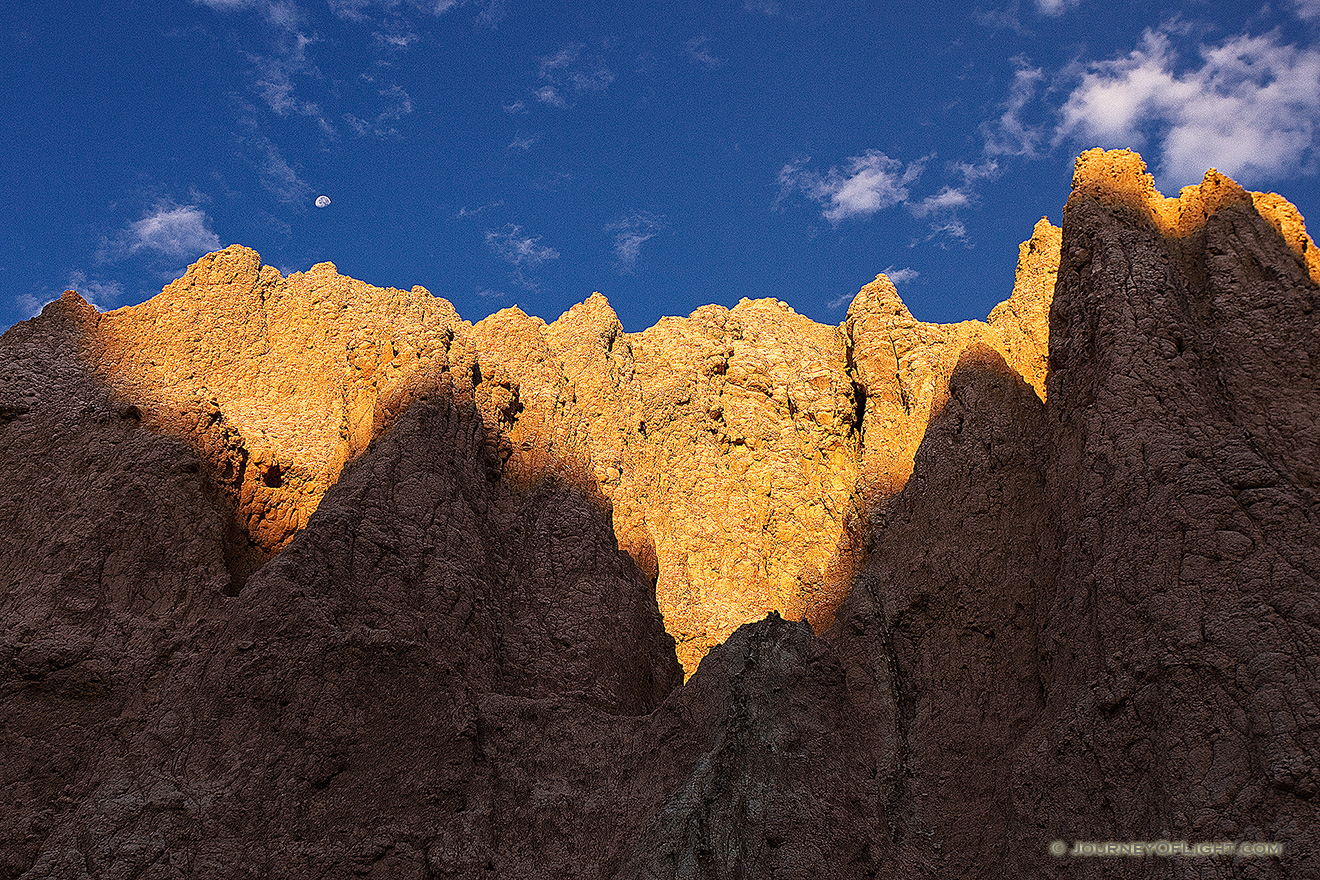 The first warm light of sunrise illuminates the erie rock formations deep in Badlands National Park as a waning moon gentle descends behind. - South Dakota Picture