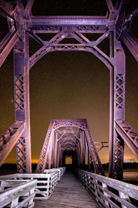 On a clear night at Niobrara State Park, I setup on the railroad trussle converted to a footbridge near the confluence of the Niobrara and Missouri Rivers.  Using a flashlight I lightpainted the bridge while exposing for the stars.  In the distance the lights of Niobrara can be seen glowing on the horizon. - Nebraska Photograph