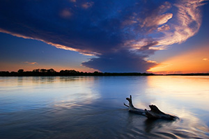 The setting sun shines through a small hole in the clouds as the Platte River flows past a lodged log.  This was taken near Schramm State Recreation Area. - Nebraska Photograph