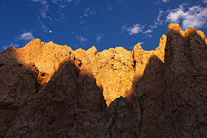 The first warm light of sunrise illuminates the erie rock formations deep in Badlands National Park as a waning moon gentle descends behind. - South Dakota Photograph