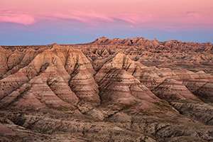 Pink clouds float above Badlands National Park while the rocky terrain below is bathed in the warm light just after sunset. - South Dakota Photograph