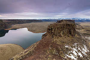 Approximately 17 million years ago lava flowed across what is now eastern Washington into Oregon.  Every few thousand years these lava flows would occur and then stop for long periods of time.  It would deposit a fine grained rock known as basalt.  After the flows stopped permanently large floods ran through this area carving out deep ravines.  Eventually, the Columbia River formed and snaked its way through Central Washington.  This landscape is the Potholes Coulee area in the Quincy Lakes unit of the Columbia Basin Wildlife Area displaying some of the unique landscape caused by these geologic events.  

I captured this image on a February morning.  It was cool, but not cold and a few remnants of a previous snow fall are still visible.  When cresting the cliff, I unfortunately startled a flock of geese in Dusty Lake below.  I took a moment to appreciate the view and then went to work capturing the beauty before me.  In the distance mountains, still covered with snow lay just beyond the unseen Columbia River.  As I was finishing up, a hawk climbed into the sky and then out of sight.  As I packed up I imagined myself as that hawk flying over the unique landscape. - Pacific Photograph