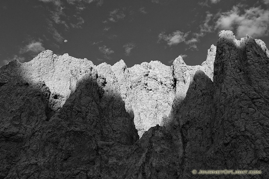 The first light of sunrise illuminates the erie rock formations deep in Badlands National Park as a waning moon gentle descends behind. - South Dakota Photography