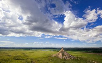 From a vantage point on a nearby bluff Chimney Rock glows in the light of the warm afternoon sun under a dark blue sky filled with clouds.