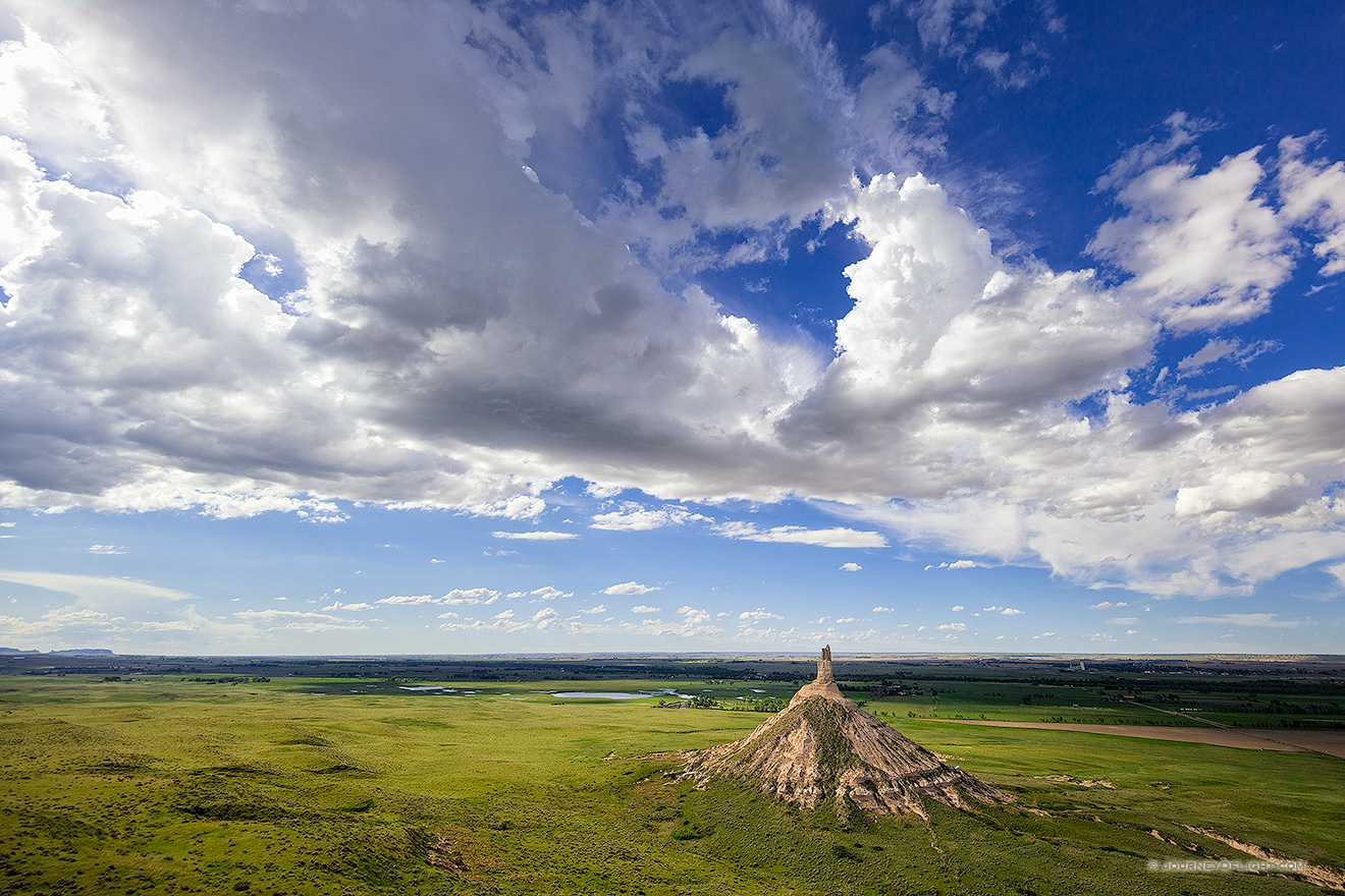 From a vantage point on a nearby bluff Chimney Rock glows in the light of the warm afternoon sun under a dark blue sky filled with clouds.