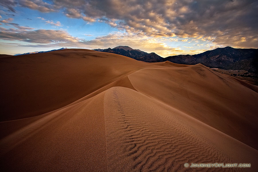 On a cool, autumn morning, silence reigned across the scenic Great Sand Dunes National Park, the quiet only occasionally broken by the sound of a breeze flowing through the dunes.  On top of one of the larger dunes, I captured the patterns of the sand as the dunes drift into one another, creating a path to the distant Sangre de Cristo Mountains.   Beyond the far peaks, the rising sun illuminates the cloud bank with a yellowish-orange tint. - Great Sand Dunes NP Photography
