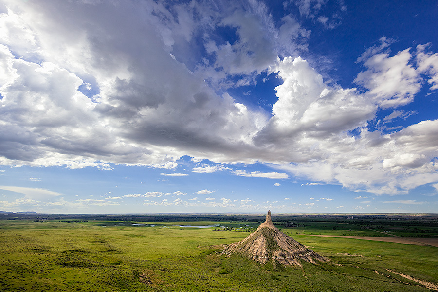 From a vantage point on a nearby bluff Chimney Rock glows in the light of the warm afternoon sun under a dark blue sky filled with clouds. - Nebraska Photography