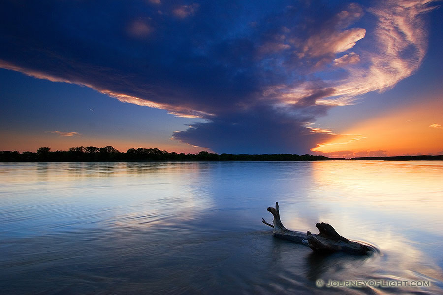 The setting sun shines through a small hole in the clouds as the Platte River flows past a lodged log.  This was taken near Schramm State Recreation Area. - Schramm SRA Photography