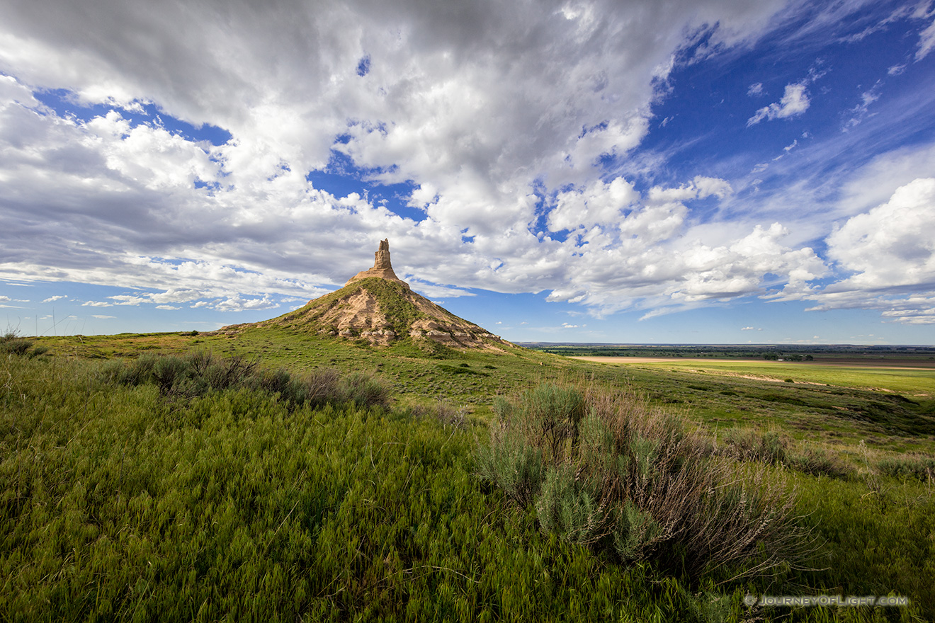 On a cool spring day, under a beautiful blue sky filled with puffy white clouds, Chimney Rock glows in the warm light of the afternoon sun. - Nebraska Picture
