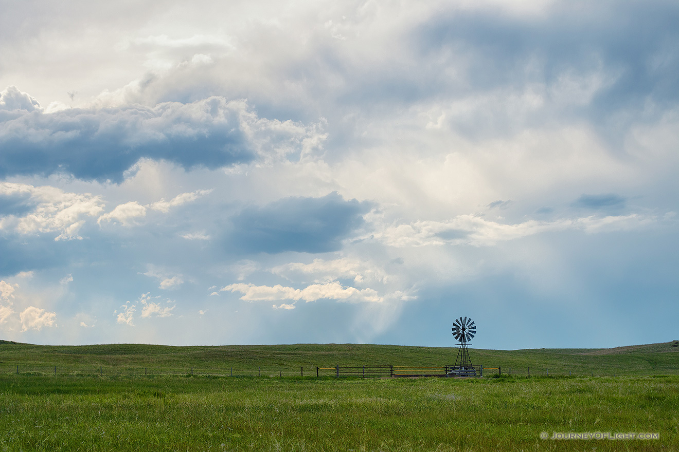 The sun peeks through a group of storm clouds illuminating a windmill deep within the Sandhills of Nebraska as storm clouds roll in the distance. - Nebraska Picture